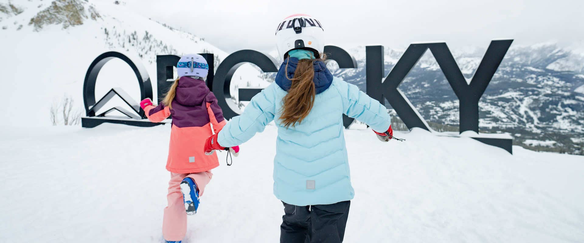Two girls in ski gear running towards a sign that says Big Sky